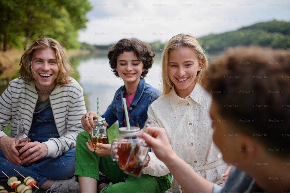 A group of young friends having fun on picnic near a lake, sitting on blanket and toasting with drinks.