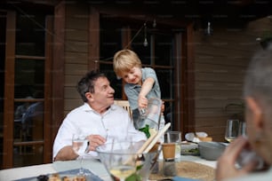 A little boy pouring lemonade to his grandfather during family celebration outside on patio.