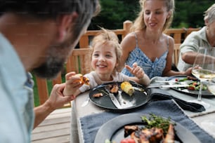 Una familia comiendo en la cena de barbacoa en el patio, niña con padres sentados a la mesa y disfrutando de la comida.