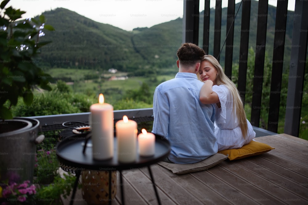 A young couple resting outdoors on terrace of tiny house in woods with view in evening, weekend away in tiny house in countryside, sustainable living.