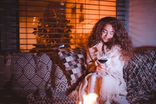 A happy young woman wrapped in blanket sitting on terrace and drinking wine in the evening.