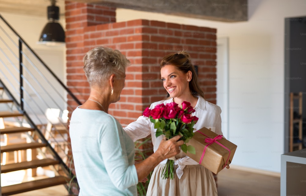 Une fille adulte heureuse apportant un cadeau et un bouquet à une mère âgée à l’intérieur à la maison.
