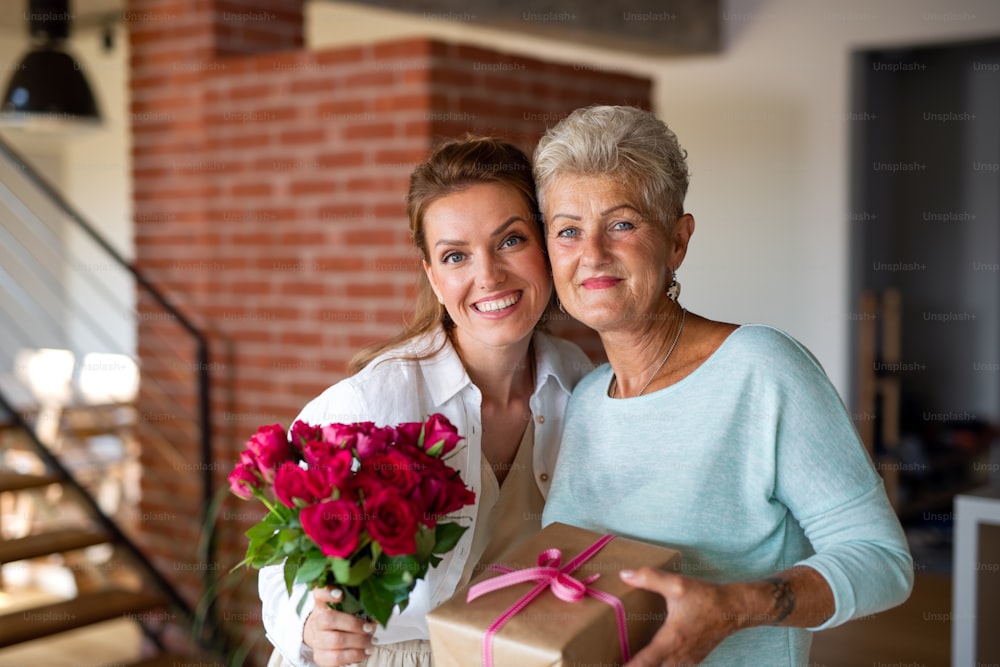 A happy senior mother getting gift and bouquet from adult daughter indoors at home, looking at camera.