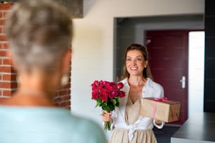 A happy adult daughter bringing gift and bouquet to senior mother indoors at home.