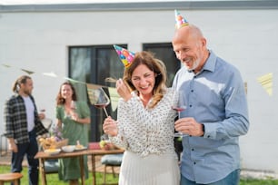 A mature couple with wine outdoors in garden at home, birthday celebration party.
