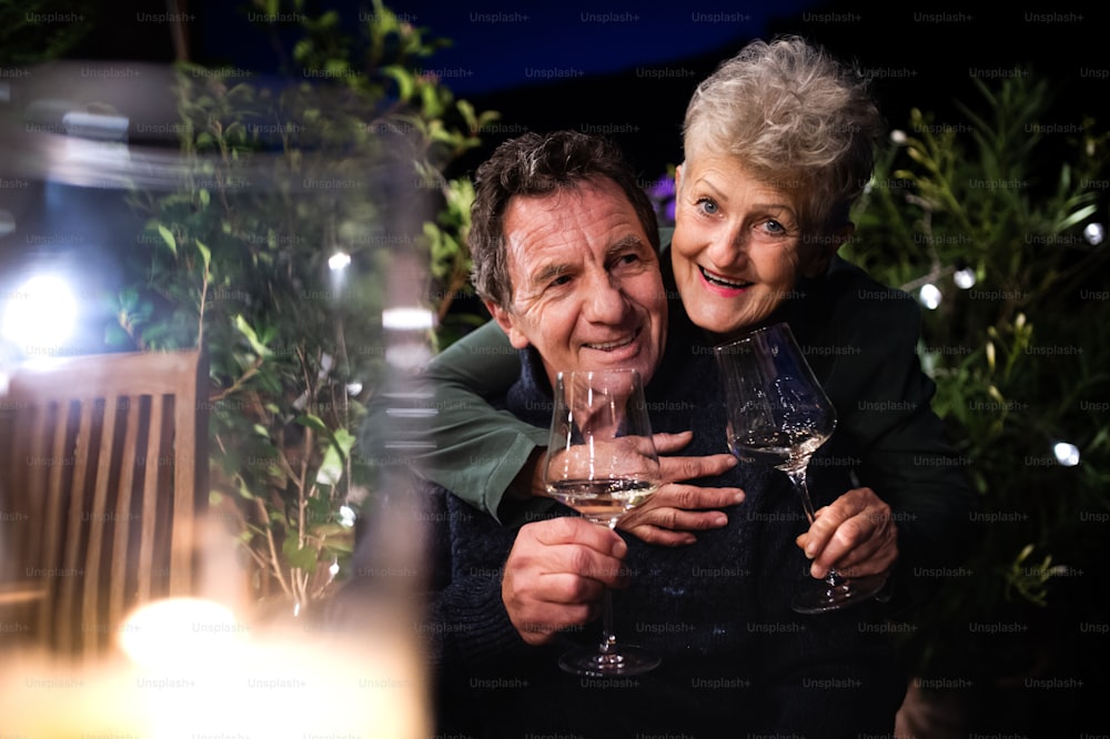 Portrait d’un couple de personnes âgées avec du vin le soir sur la terrasse, regardant la caméra.