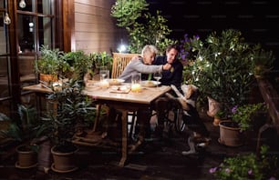 Feliz pareja mayor en silla de ruedas cenando por la noche en la terraza, alimentando perro.