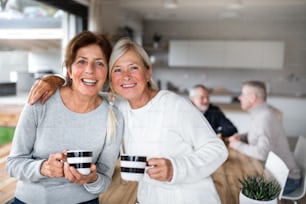 Front view portrait of senior women friends with coffee at home, looking at camera.