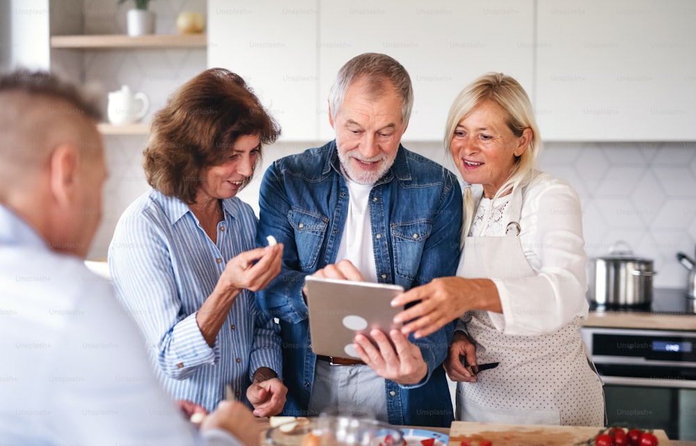 Vista frontal do grupo de alegres amigos seniores com tablet no jantar em casa, cozinhando.