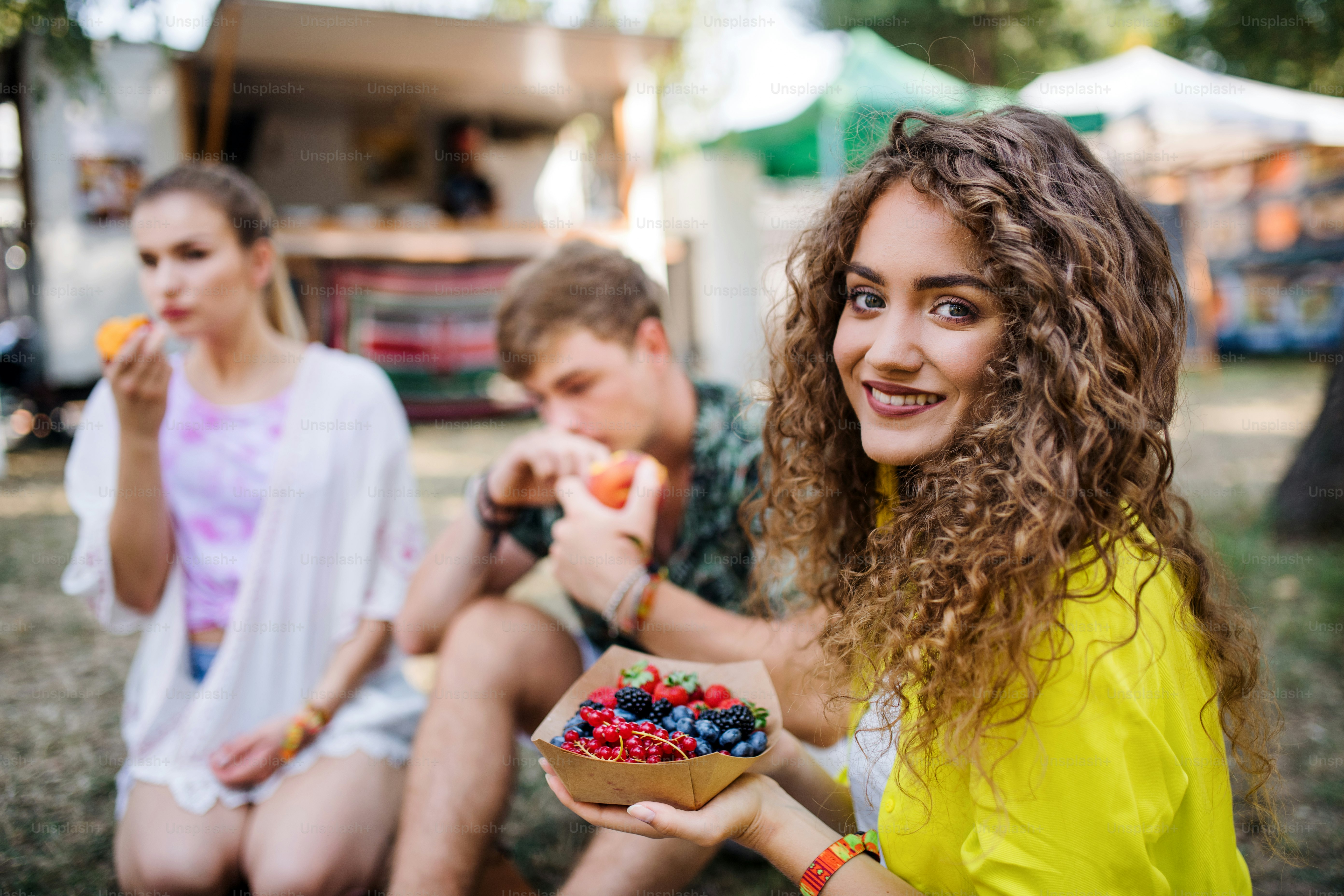 Beautiful young girl with friends sitting on ground at summer festival, eating fruit.