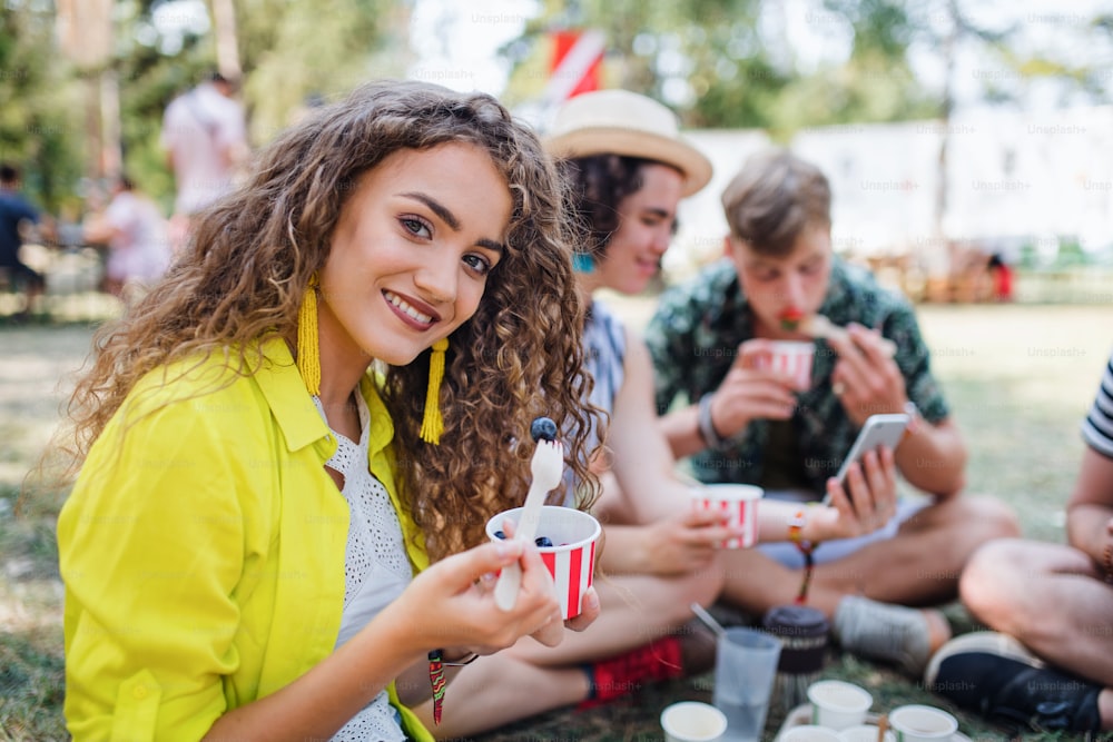 Beautiful young girl with friends sitting on ground at summer festival, eating fruit.