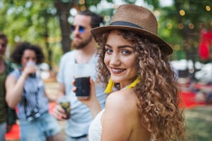 Beautiful young girl with drink and friends standing at summer festival.