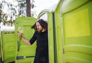 Young man by mobile toilet at summer festival. Copy space.