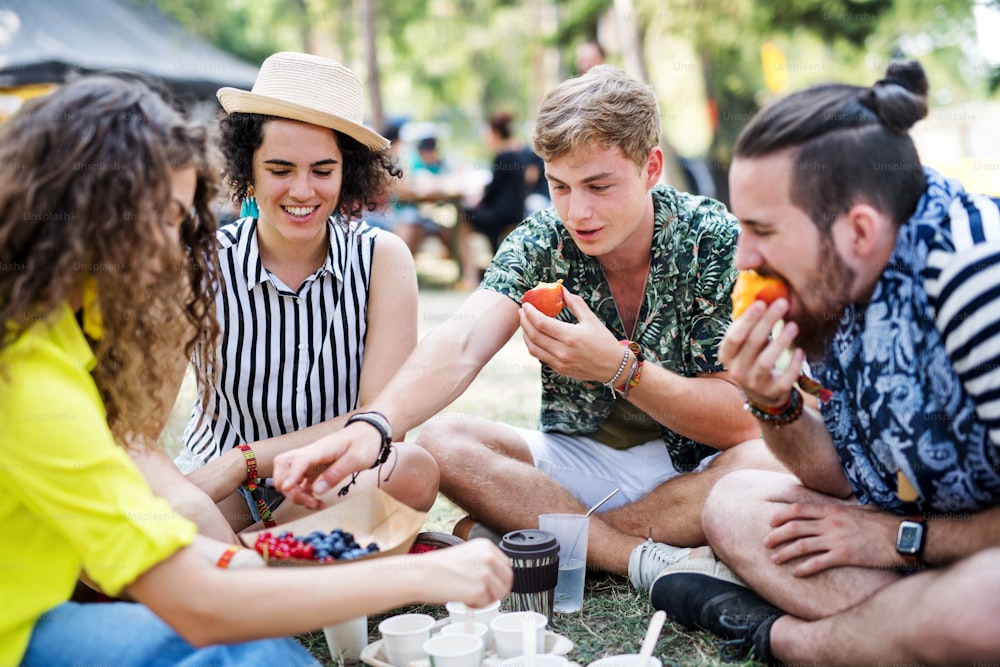 Groupe de jeunes amis joyeux au festival d’été, assis par terre et mangeant.
