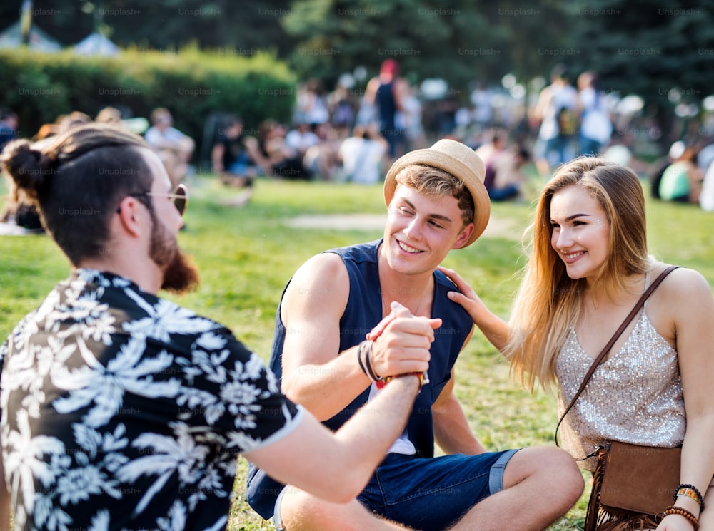 Gruppo di giovani amici allegri seduti a terra al festival estivo, stringendo la mano.