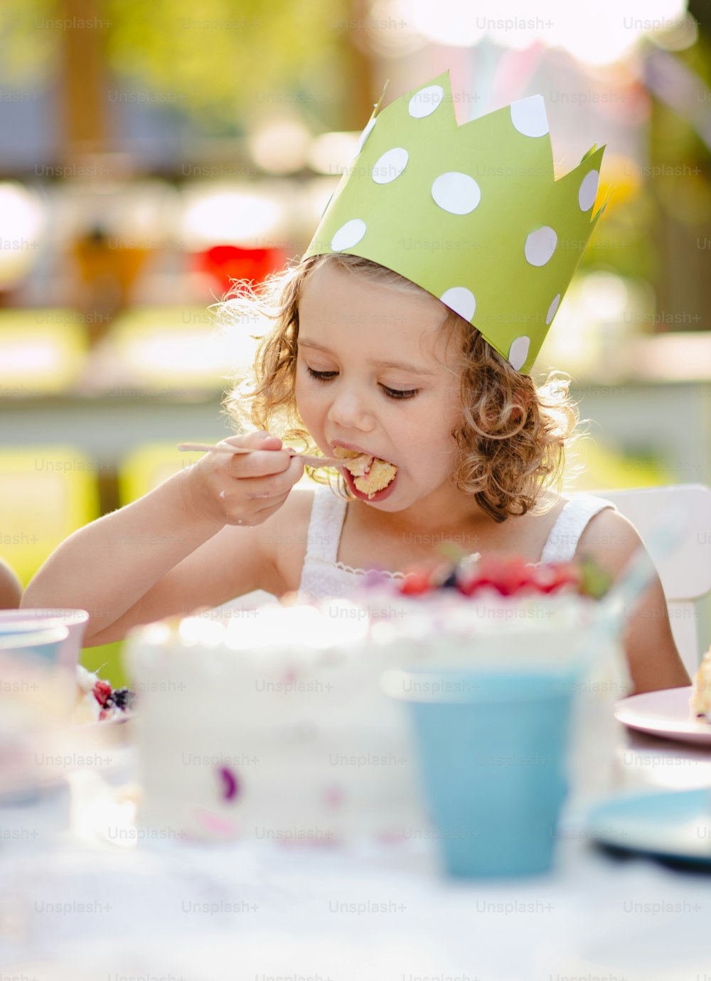 Kleines Mädchen mit Partyhut, das im Sommer draußen im Garten sitzt und Kuchen isst. Ein Festkonzept.