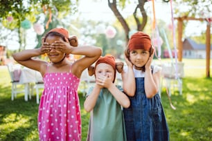 Front view of small children with masks outdoors on garden party in summer, playing.