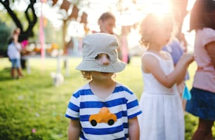 Niño pequeño con amigos parados al aire libre en una fiesta en el jardín en verano, con un agujero en su sombrero. Concepto de celebración.