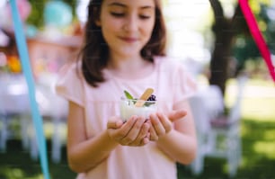 Front view of small girl outdoors in garden in summer, holding dessert in cup. A celebration concept.