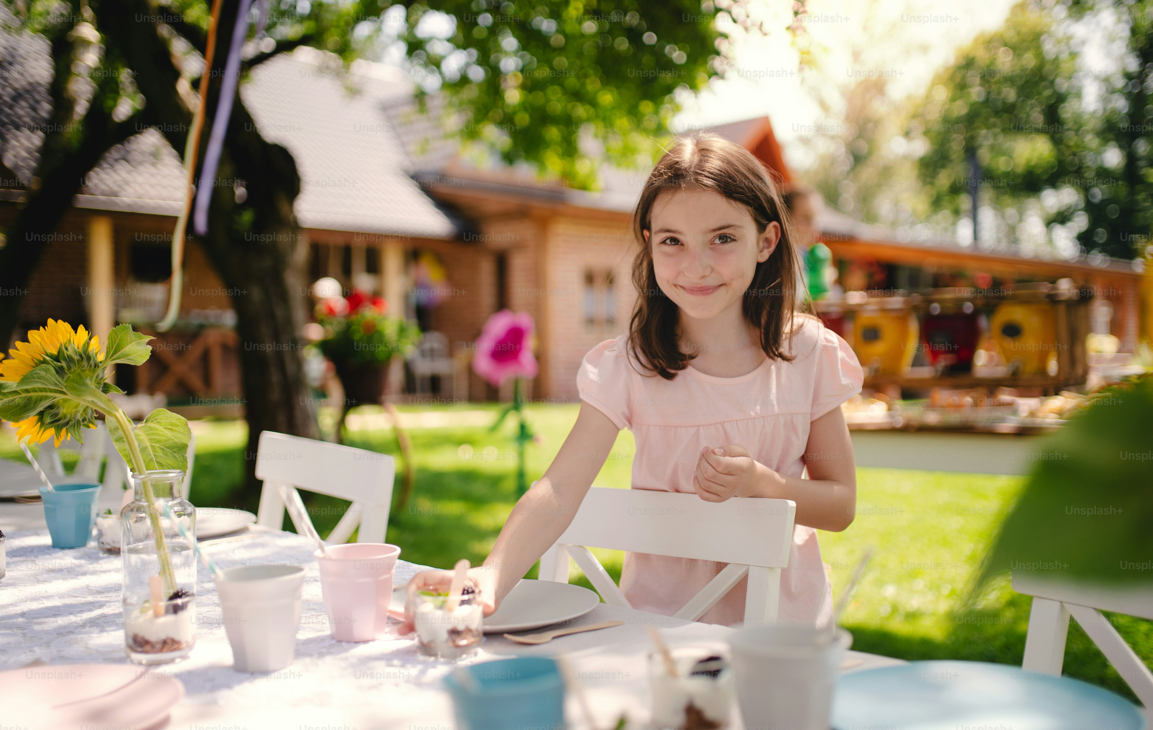 Portrait of small girl standing by table outdoors in garden in summer on party, looking at camera.