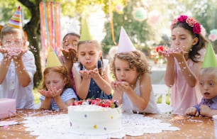 A portrait of children with cake standing around table on birthday party in garden in summer.