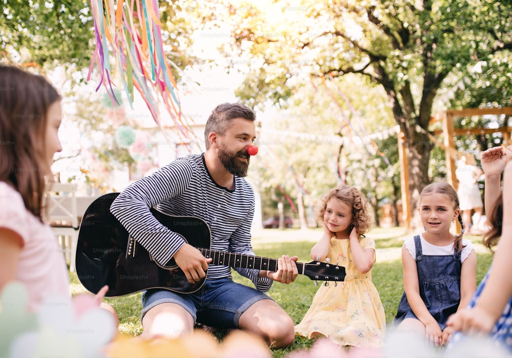 Un homme avec de jeunes enfants assis par terre à l’extérieur dans le jardin en été, jouant de la guitare.
