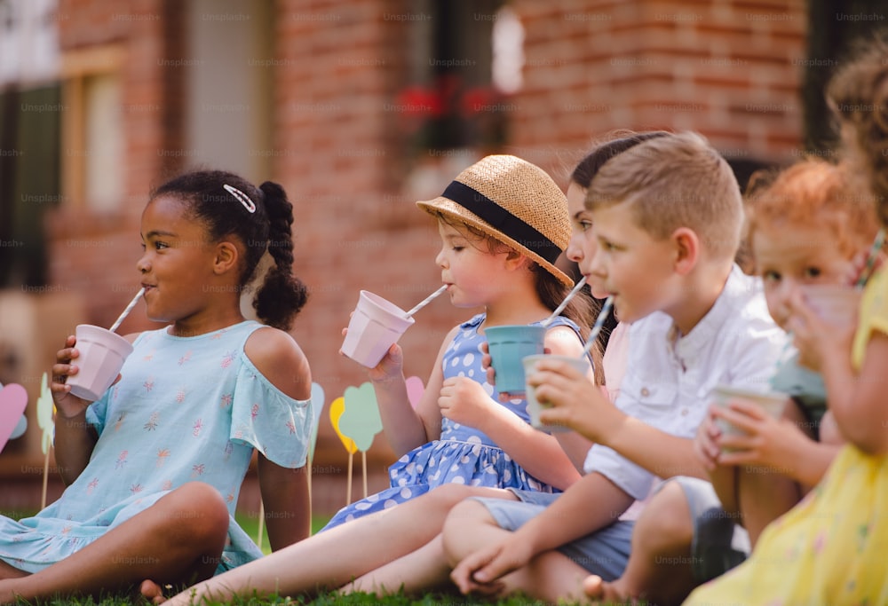 Bambini piccoli seduti a terra all'aperto in giardino in estate, bevendo. Un concetto di celebrazione.