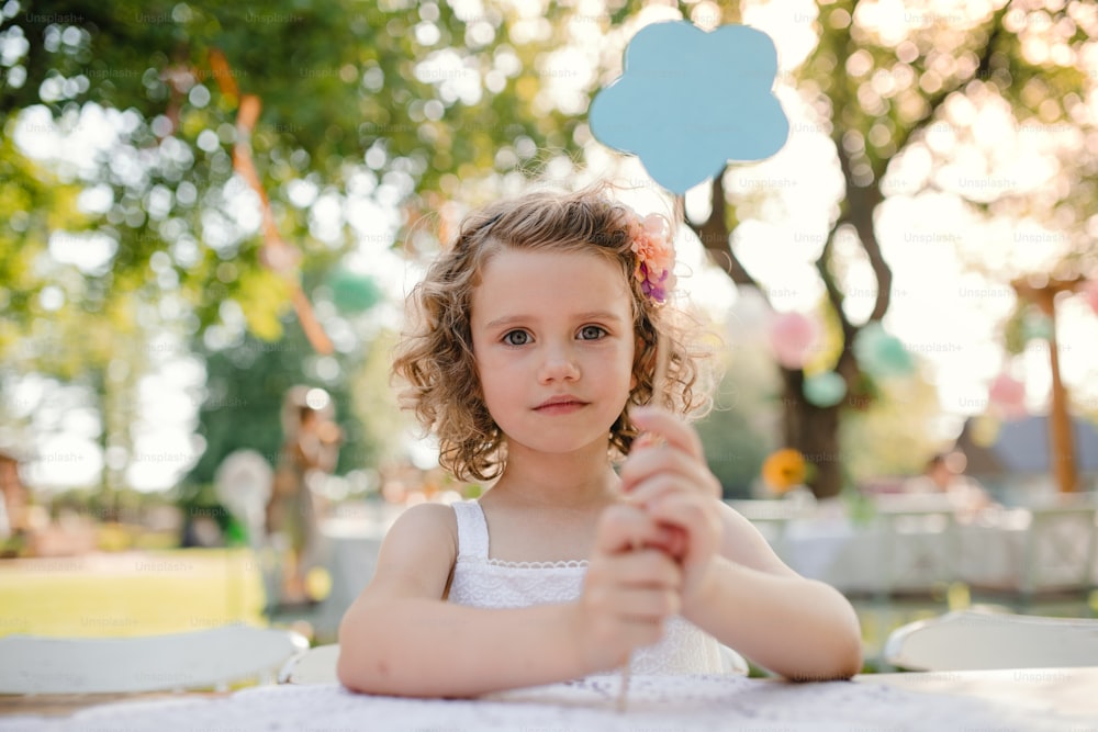 Small girl sitting outdoors in garden in summer, a birthday celebration concept.