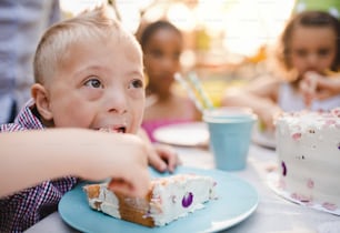 Down syndrome child with friends on birthday party outdoors in garden, eating cake.