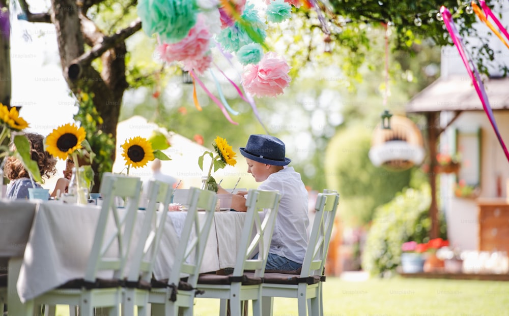 A small boy sitting at the table outdoors on garden party in summer, eating.