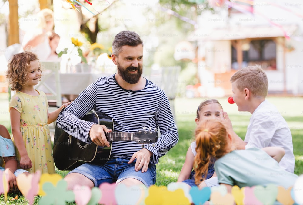 Un hombre con niños pequeños sentado en el suelo al aire libre en el jardín en verano, tocando la guitarra.