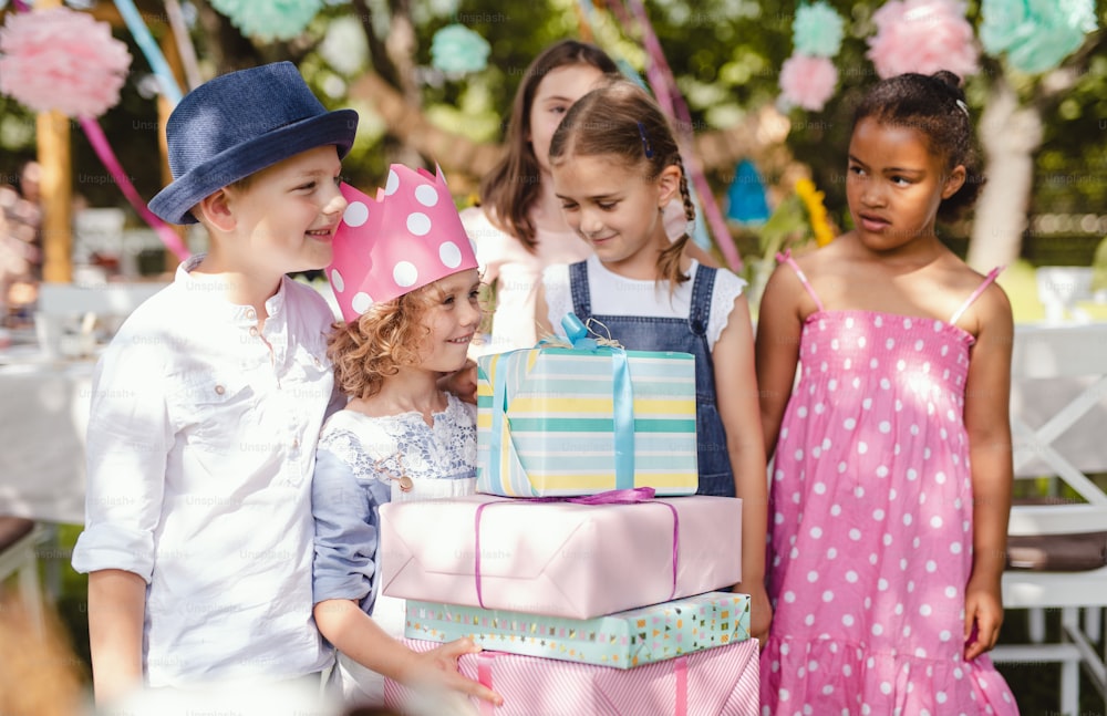 A portrait of small girl with friends and presents outdoors in garden in summer.