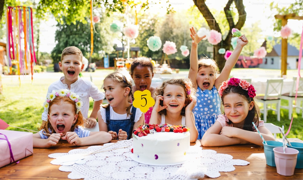 A portrait of children with cake standing around table on birthday party in garden in summer.
