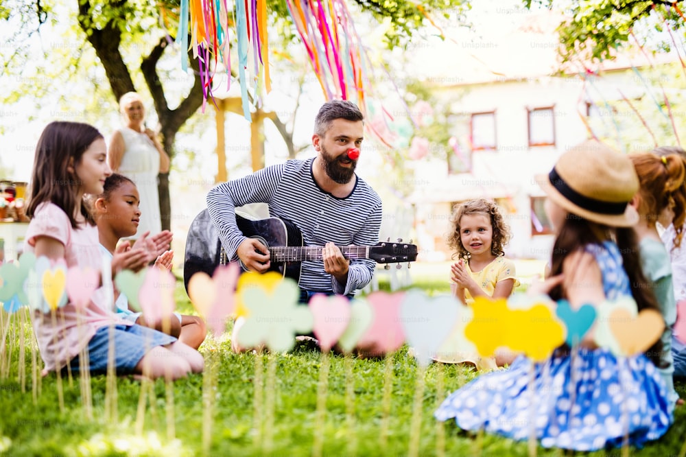 Un hombre con niños pequeños sentado en el suelo al aire libre en el jardín en verano, tocando la guitarra.