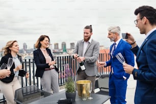A large group of joyful businesspeople having a party outdoors on roof terrace in city, opening bottle of champagne.