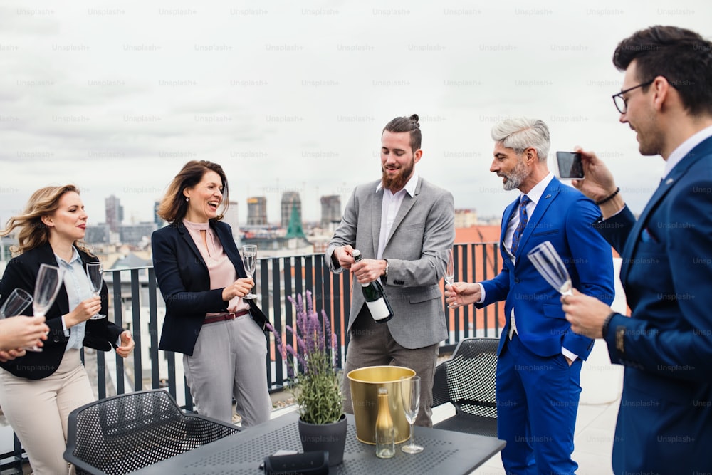 A large group of joyful businesspeople having a party outdoors on roof terrace in city, opening bottle of champagne.