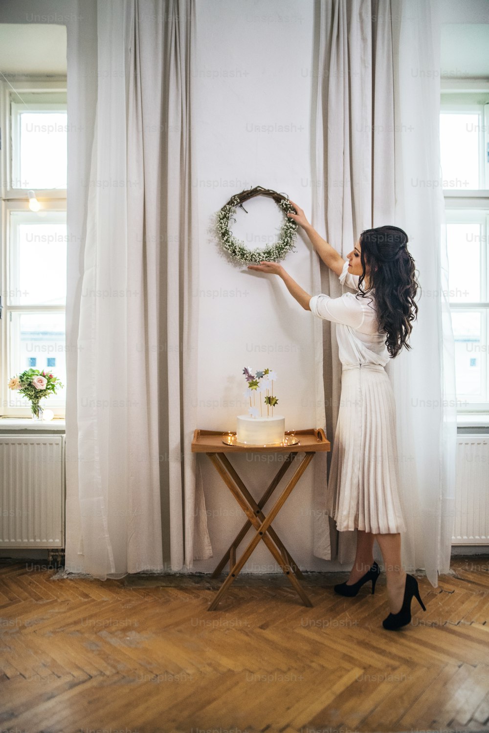 A woman hanging up a wreath on wall on indoor party, a cake on stand. Copy space.