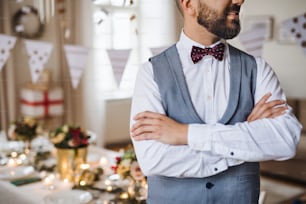 Una parte centrale di un uomo maturo hipster in piedi in una stanza allestita per una festa, le braccia incrociate.