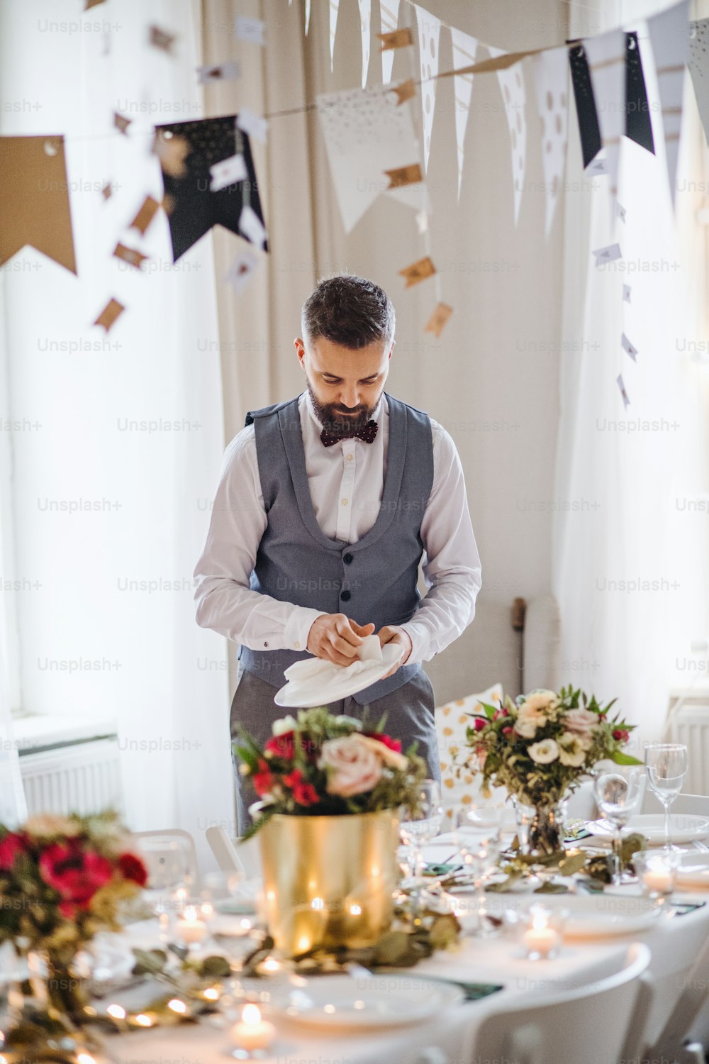 A handsome mature man indoors in a room set for a party, polishing plates.