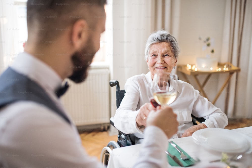 Une femme âgée en fauteuil roulant assise à la table lors d’une fête d’anniversaire à l’intérieur, faisant claquer des verres.