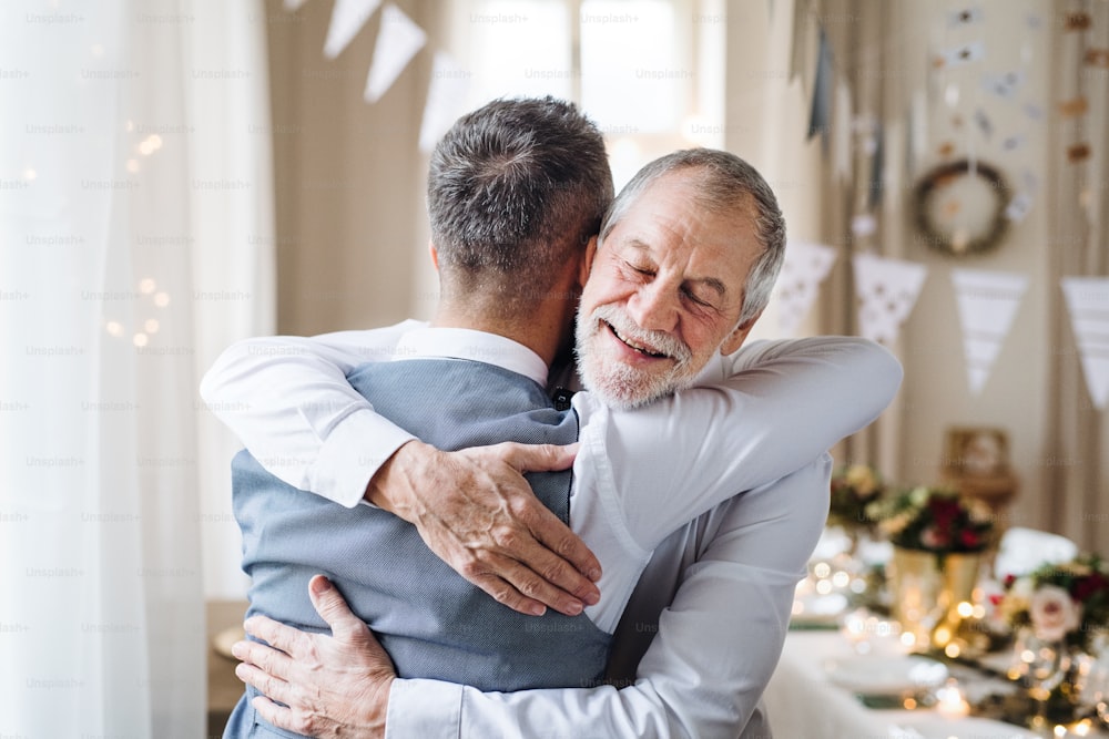 A portrait of a senior and hipster mature man standing indoors in a room set for a party, hugging.