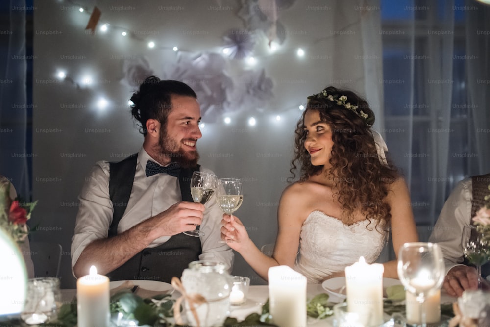 A young happy couple sitting at a table on a wedding, clinking glasses.