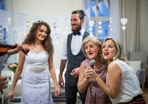 A young cheerful bride and groom with other guests dancing and singing on a wedding reception.