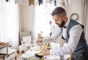 Un uomo maturo con fiocco e gilet che prepara un tavolo per una festa al coperto, lucidando un bicchiere.