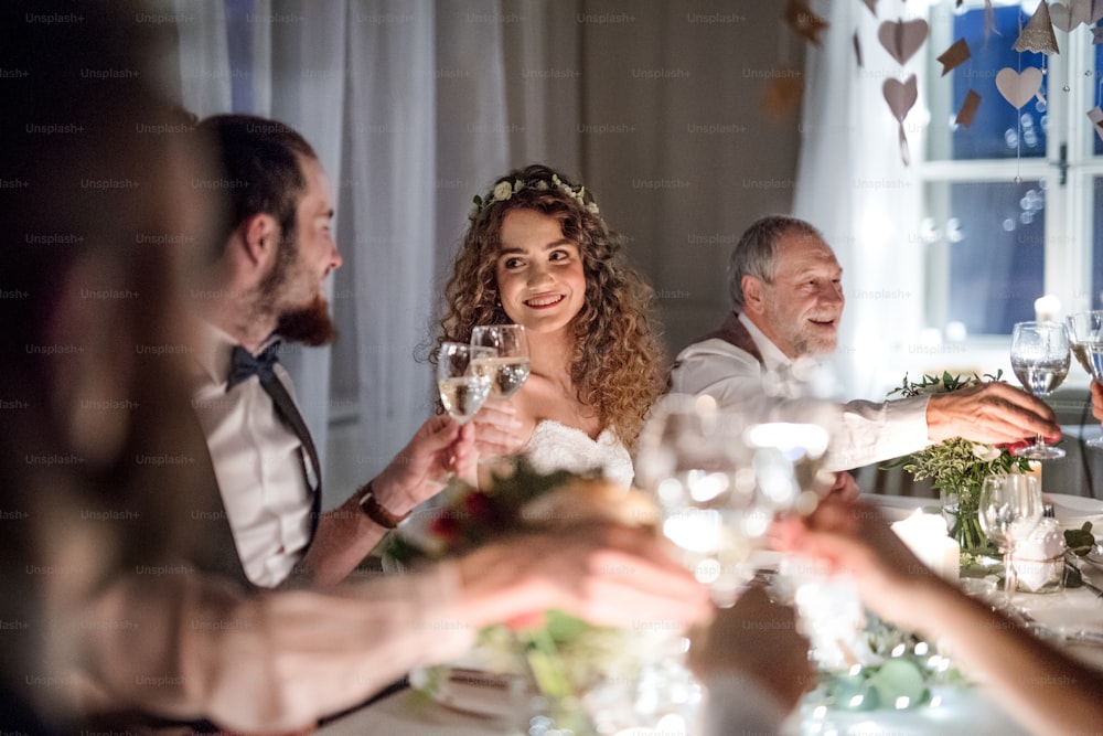 Uma noiva e um noivo e outros convidados sentados em uma mesa em um casamento, piscando os copos.
