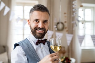 A portrait of a mature man standing indoors in a room set for a party, holding a glass of wine. Copy space.