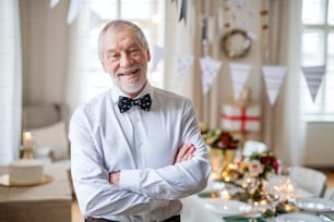 A portrait of a senior man standing indoors in a room set for a party, arms crossed. Copy space.