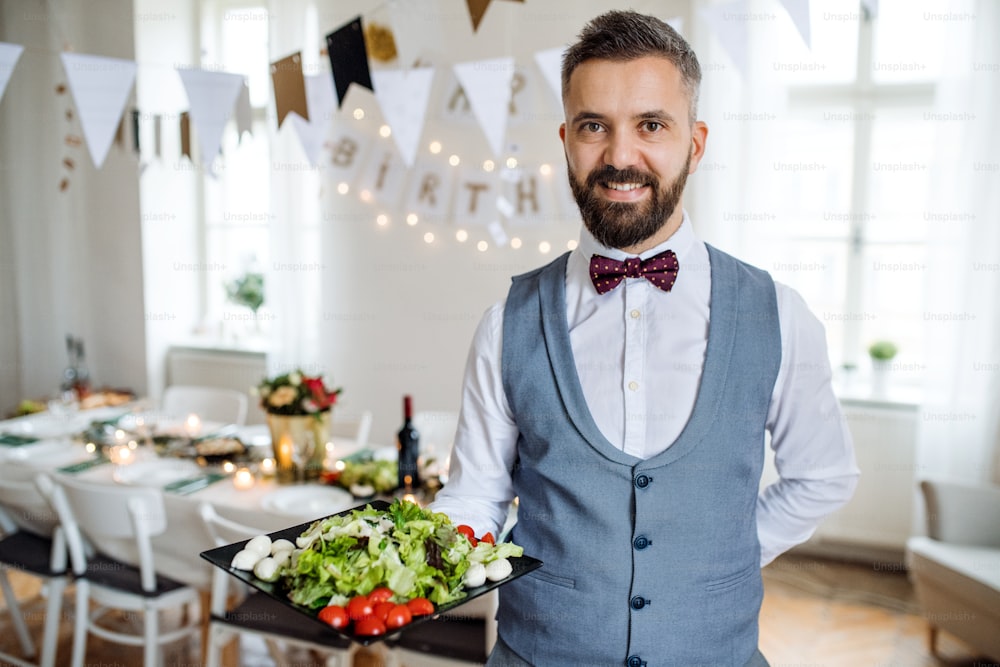 Un homme mûr debout à l’intérieur dans une pièce aménagée pour une fête, tenant un plateau avec des légumes.