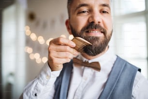 Hipster man standing indoors in a room set for a party, combing beard with a comb.