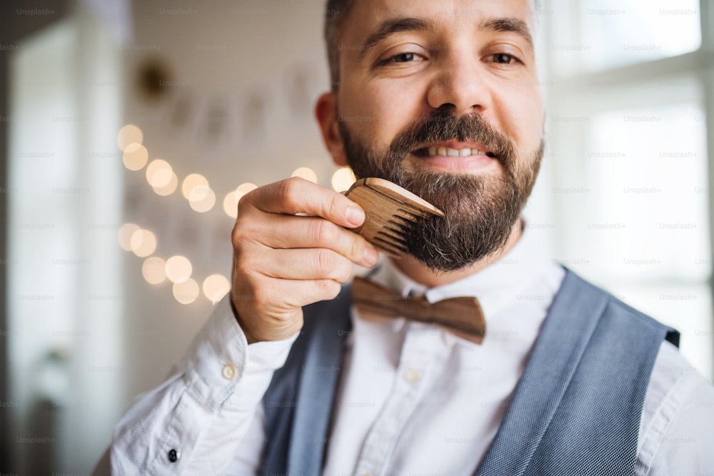Homme hipster debout à l’intérieur dans une pièce aménagée pour une fête, peignant la barbe avec un peigne.